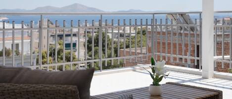 A balcony with wicker furniture, a potted plant, and a view of the sea and mountains.