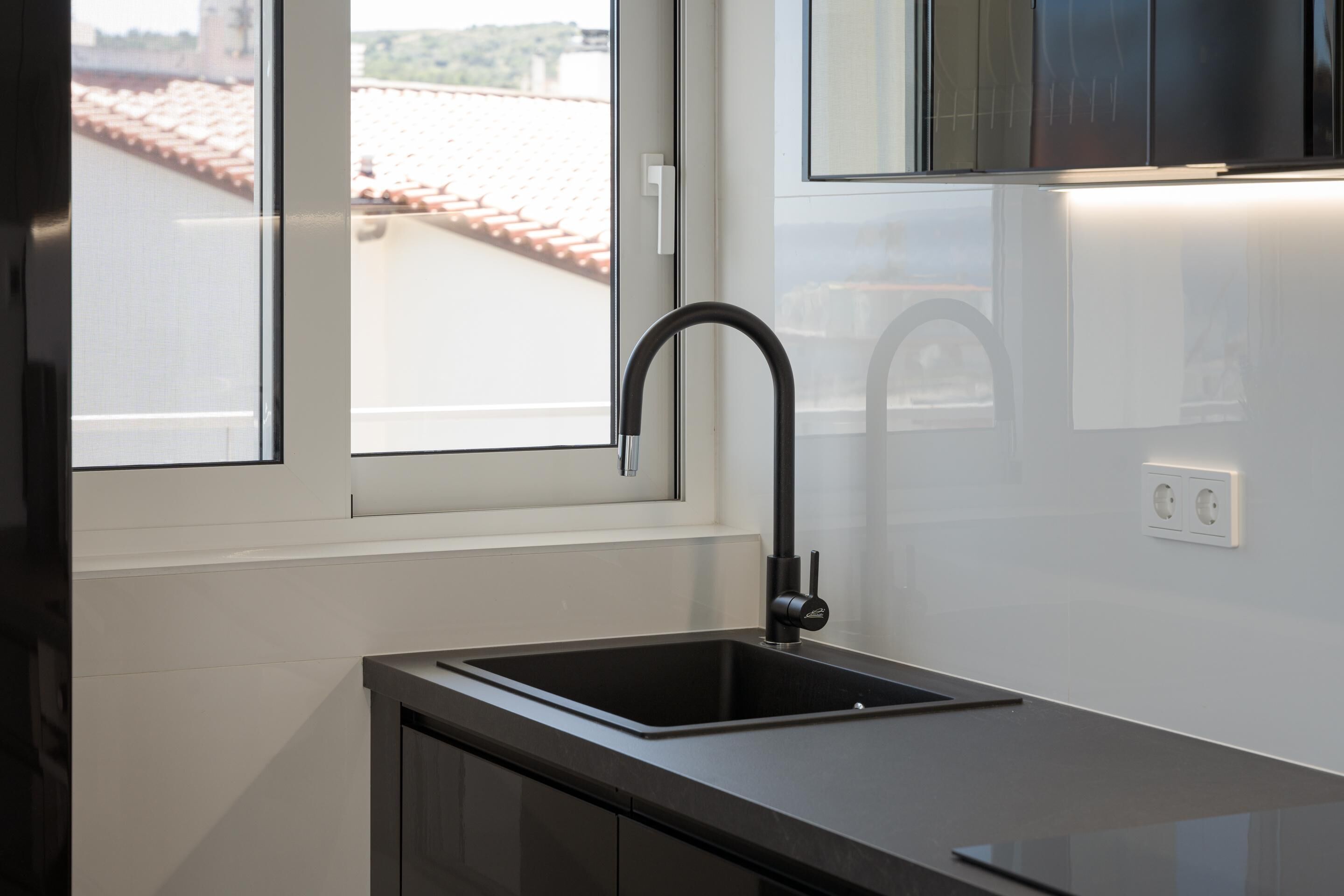 A modern kitchen with a black faucet, white tiles, and a window overlooking a red-tiled roof.