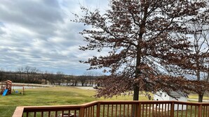 A wooden deck with a railing, a playground with a slide, and a tree in the background. - Enjoy this spacious ranch-style house next to the quilting capital of the world. (Hamilton)