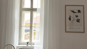 Une pièce avec une table blanche, une chaise, un radiateur et une fenêtre avec des rideaux blancs. Un tableau encadré est accroché au mur.