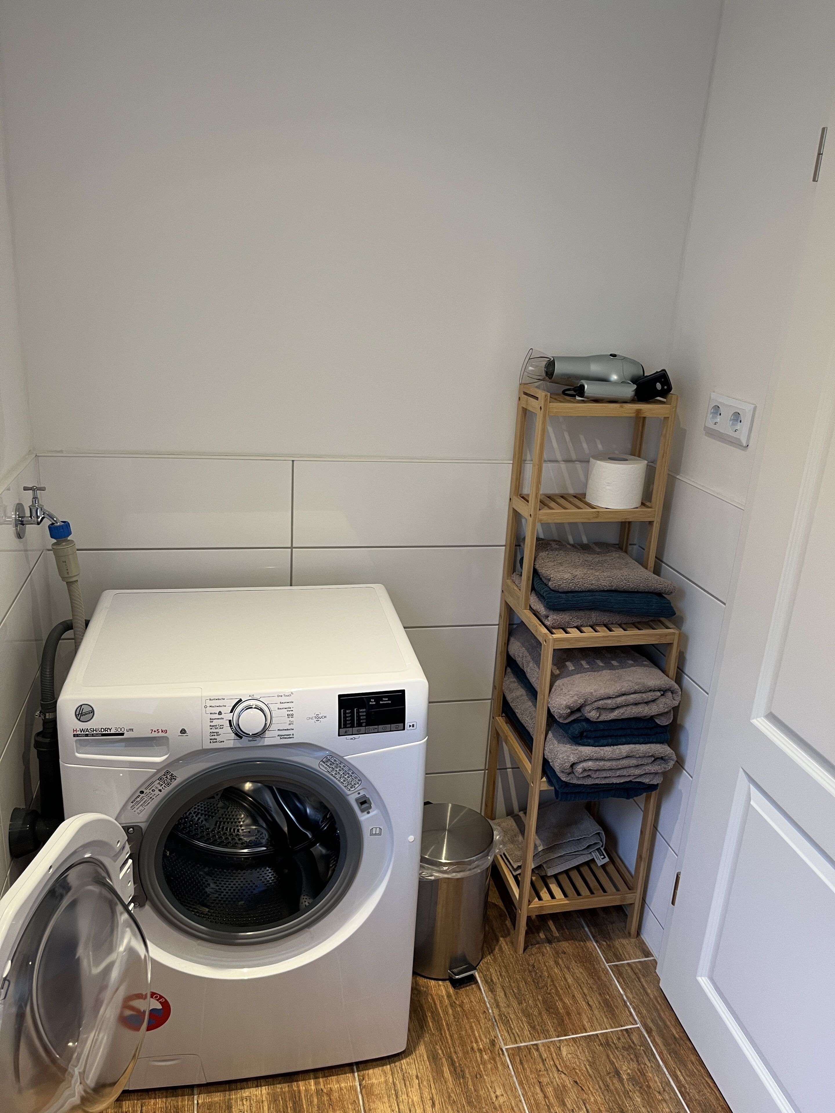 Une petite buanderie équipée d’un lave-linge à chargement frontal, d’une poubelle en métal et d’une étagère en bois où sont rangées les serviettes.