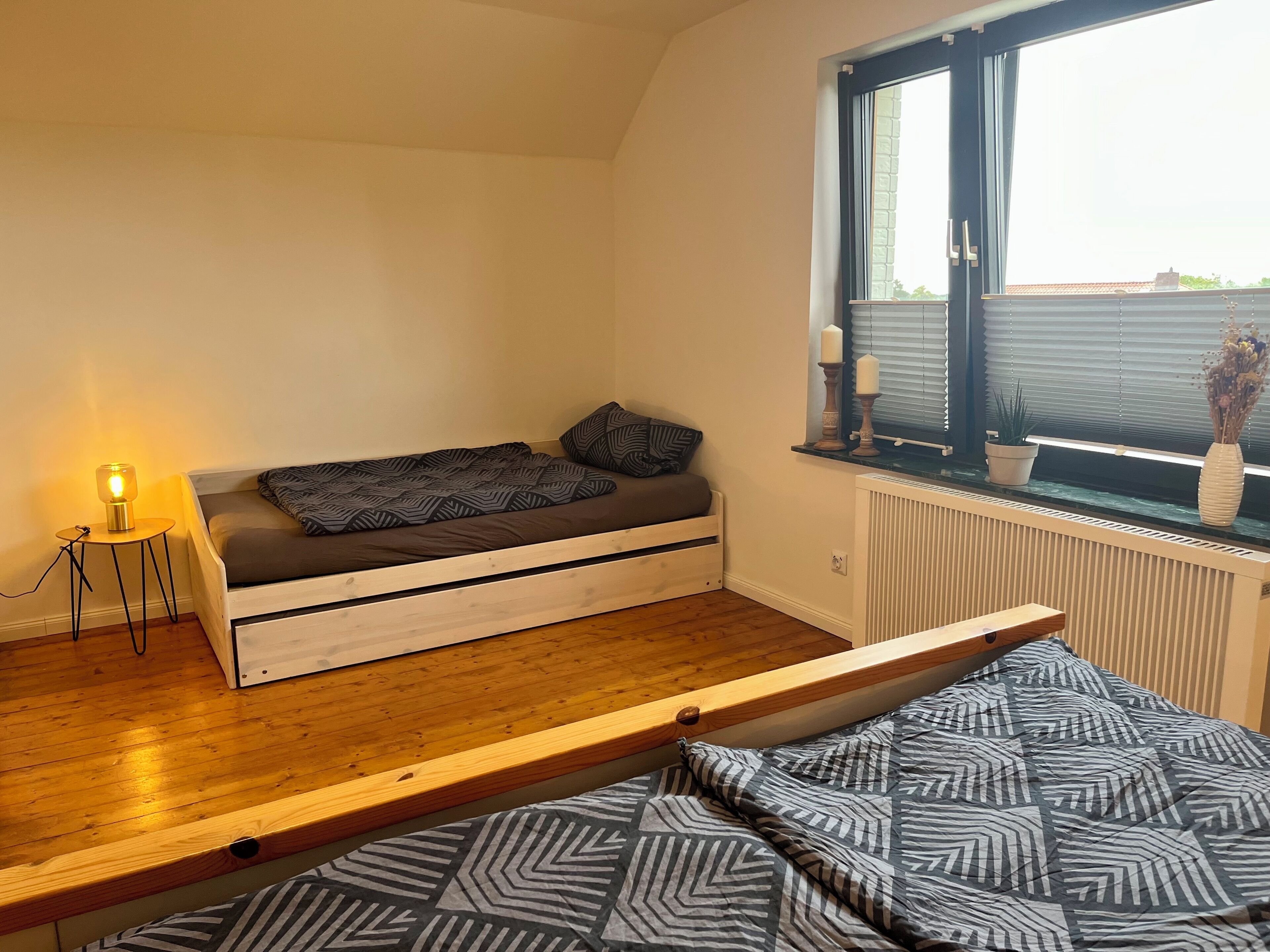 Ein kleines Zimmer mit Schlafkoje, einem kleinen Nachttisch mit Lampe, einem Heizkörper und einem Fenster mit Jalousien.