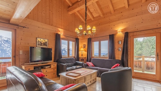 A cozy log cabin interior with a television, leather sofas, a wooden coffee table, and large windows offering mountain views.