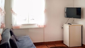 Una habitación pequeña con una cama, un radiador, un televisor sobre un mueble y una ventana con cortinas translúcidas.