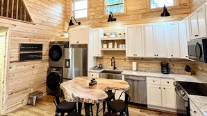 A cozy kitchen with wooden walls, a round wooden table, and bar stools. There are built-in appliances, white cabinets, and a window. - New Modern A-Frame Near Mammoth Cave National Park with Nolin Lake Views! (Cub Run)