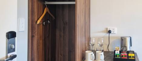 A wooden wardrobe with a hanging rod, a door with a sign instructing to make up the room, and a countertop with two white mugs, two wine glasses, and a coffee maker.