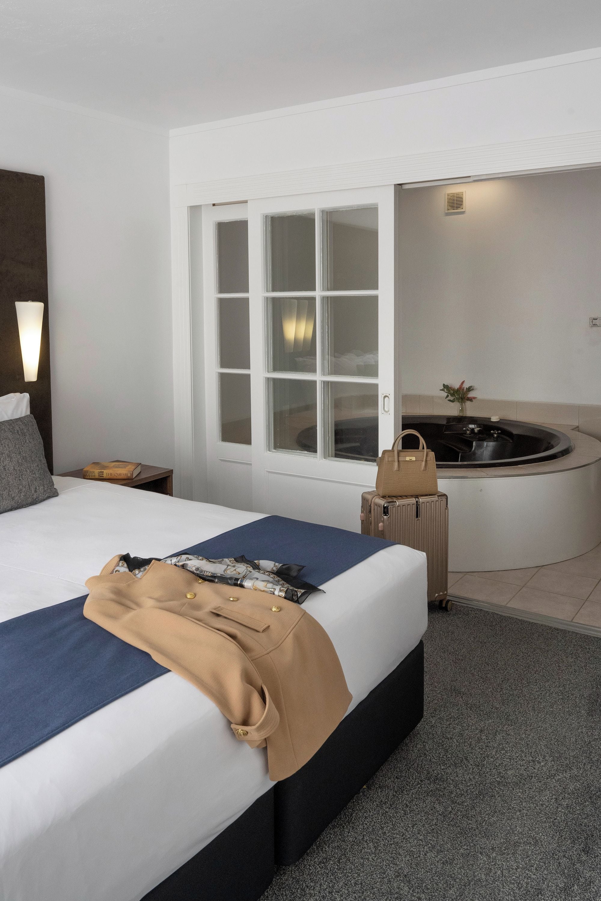 A hotel room with a bed, a window, a wall-mounted light, a bedside table, a suitcase, and a bathtub.