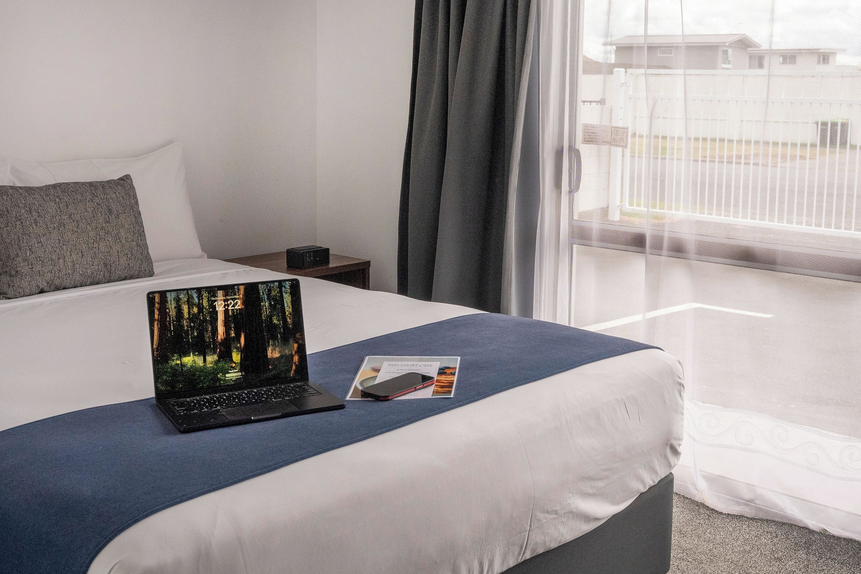 Une chambre d’hôtel avec un lit, un ordinateur portable posé sur le lit, une table de chevet et une fenêtre avec des rideaux.