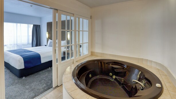 A hotel room with a large bathtub, a bed with a blue accent, and a view through a glass door.