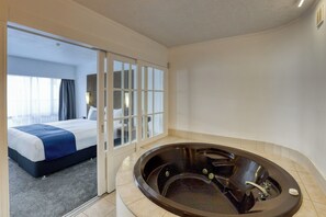 A hotel room with a large bathtub, a bed with a blue accent, and a view through a glass door.