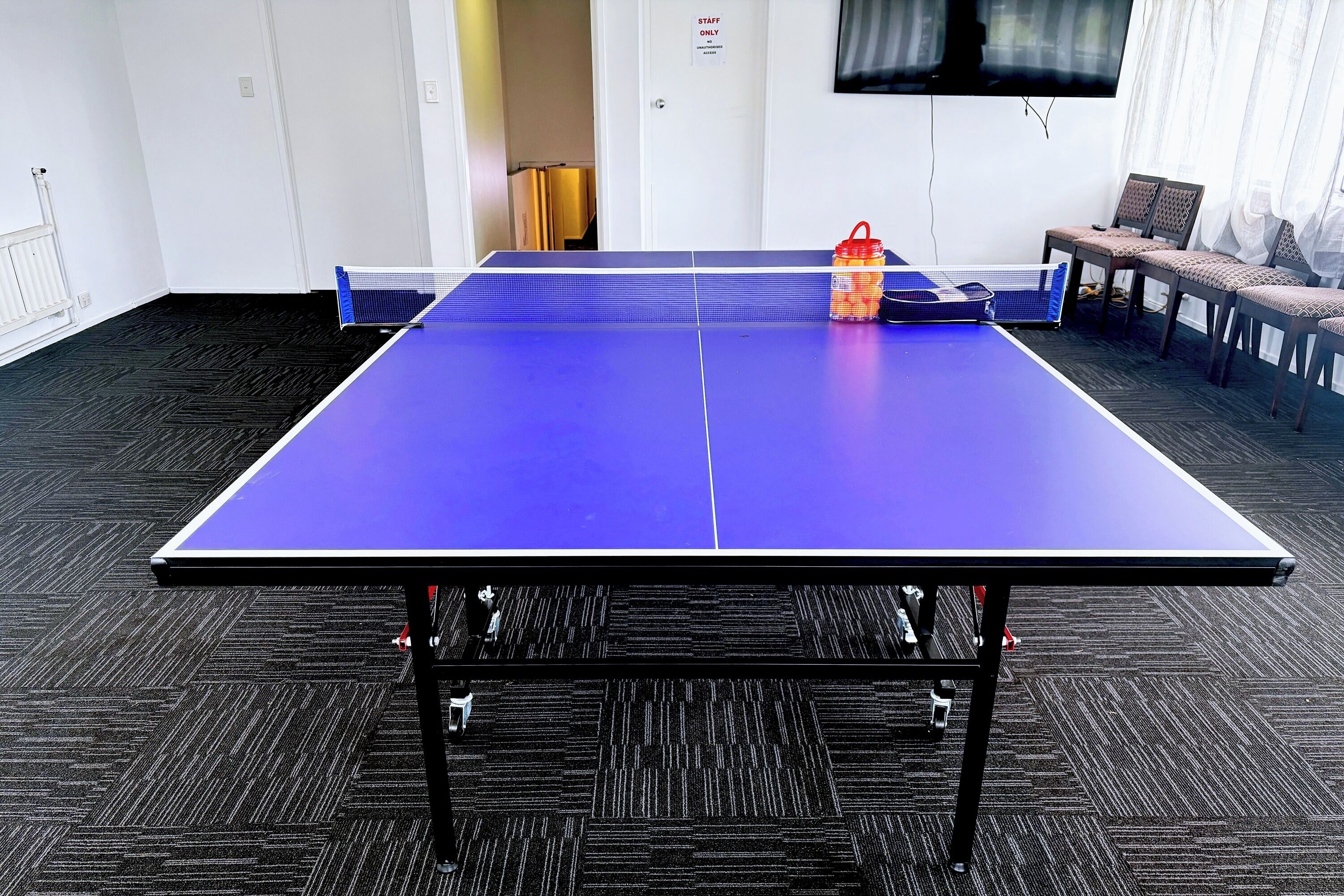 A ping pong table in a room with a carpeted floor, a TV on the wall, and a door marked 'STAFF ONLY'.