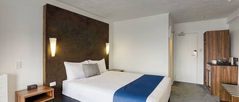 A modern hotel room with a large bed, bedside tables, a wooden headboard, and a small kitchenette area.