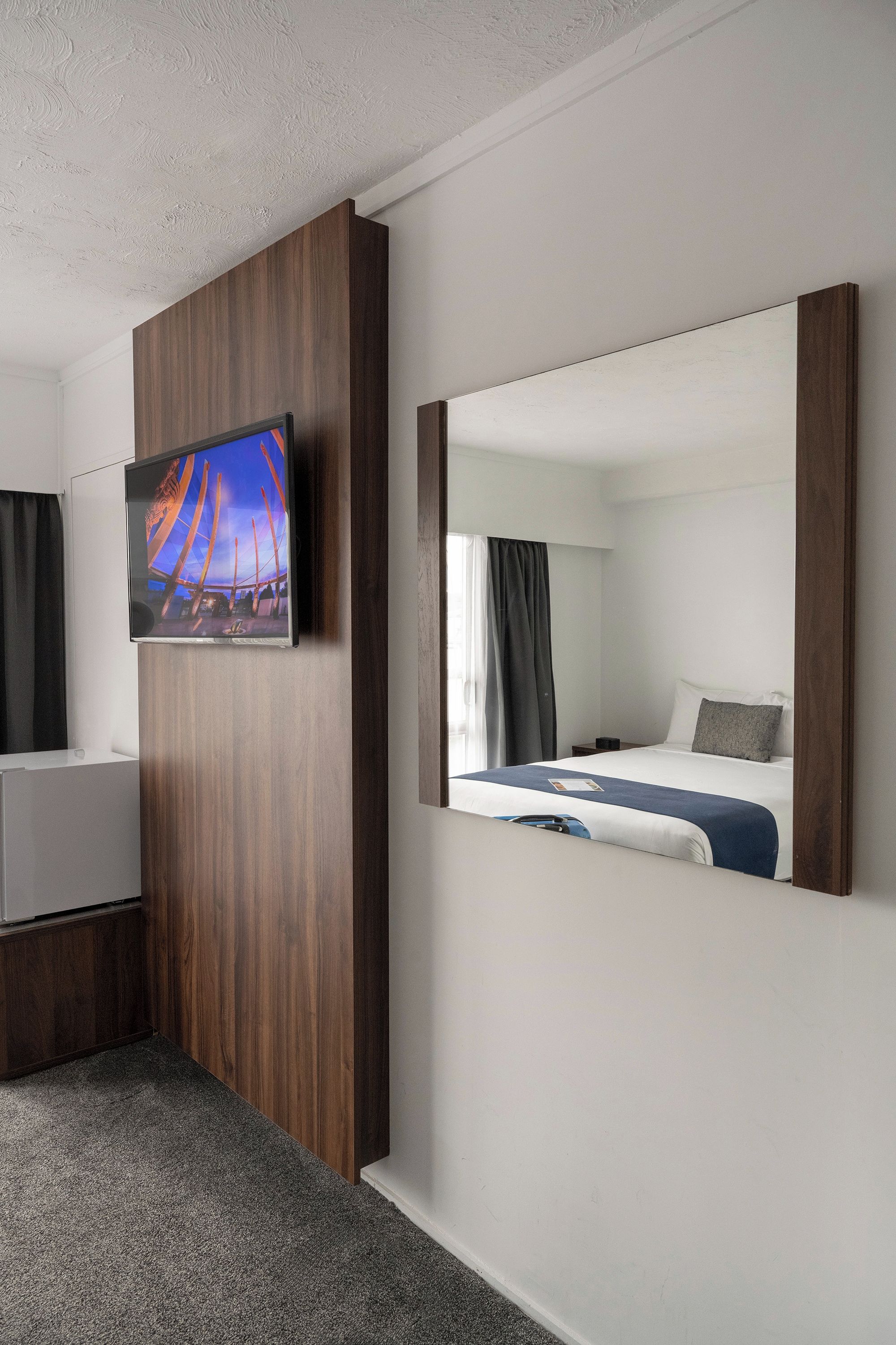 A hotel room with a bed, a television mounted on a wooden panel, and a large mirror reflecting the room.