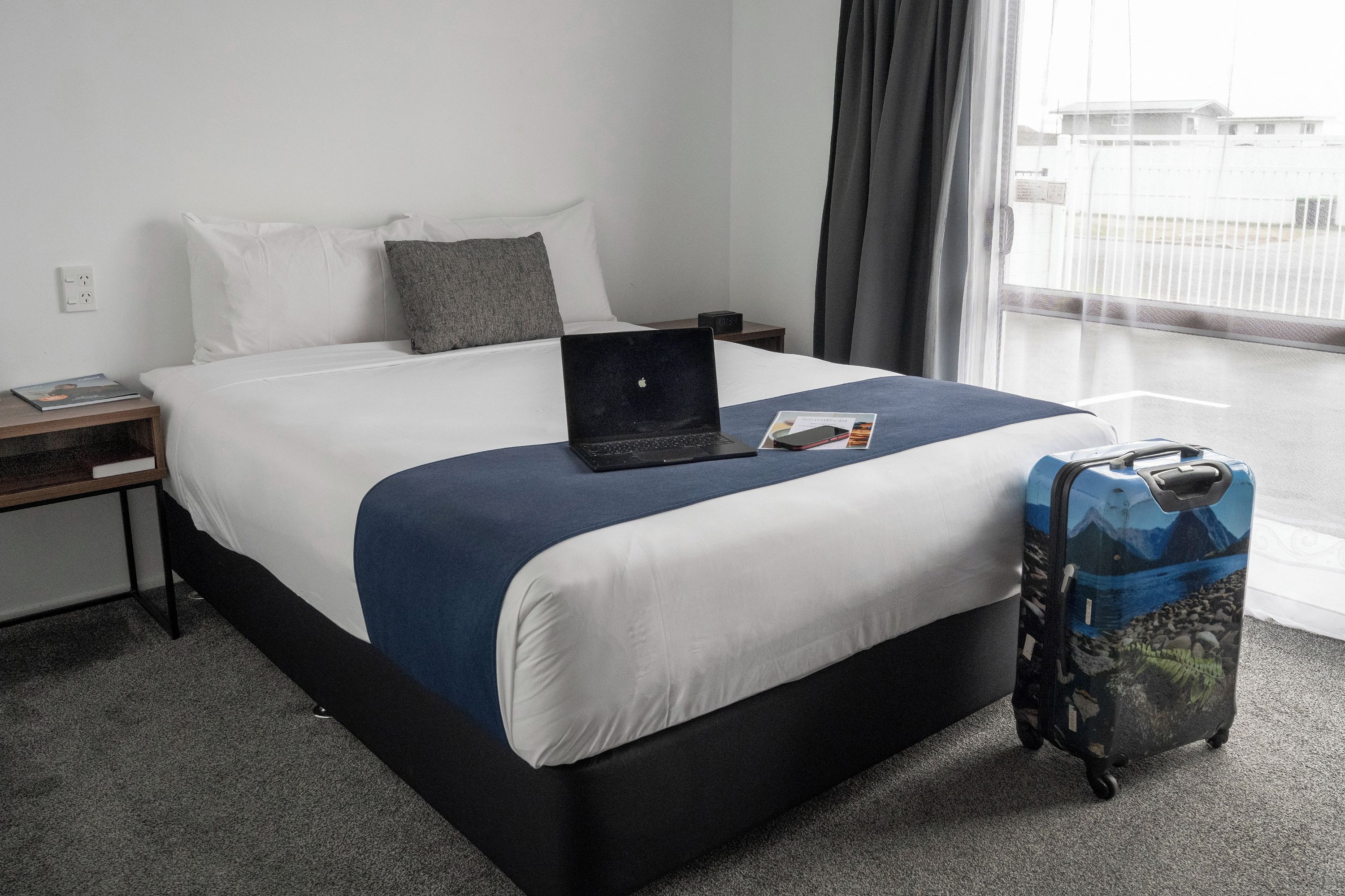 Habitación de hotel con una cama, una computadora portátil, una maleta y una ventana con vistas a edificios.
