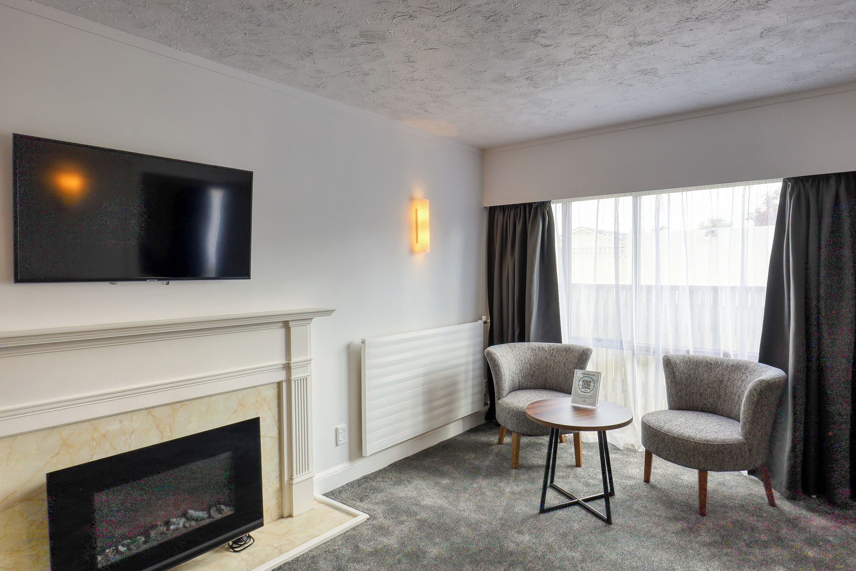 A modern living room with a fireplace, a flat-screen TV, two armchairs, a small table, and a large window with curtains.