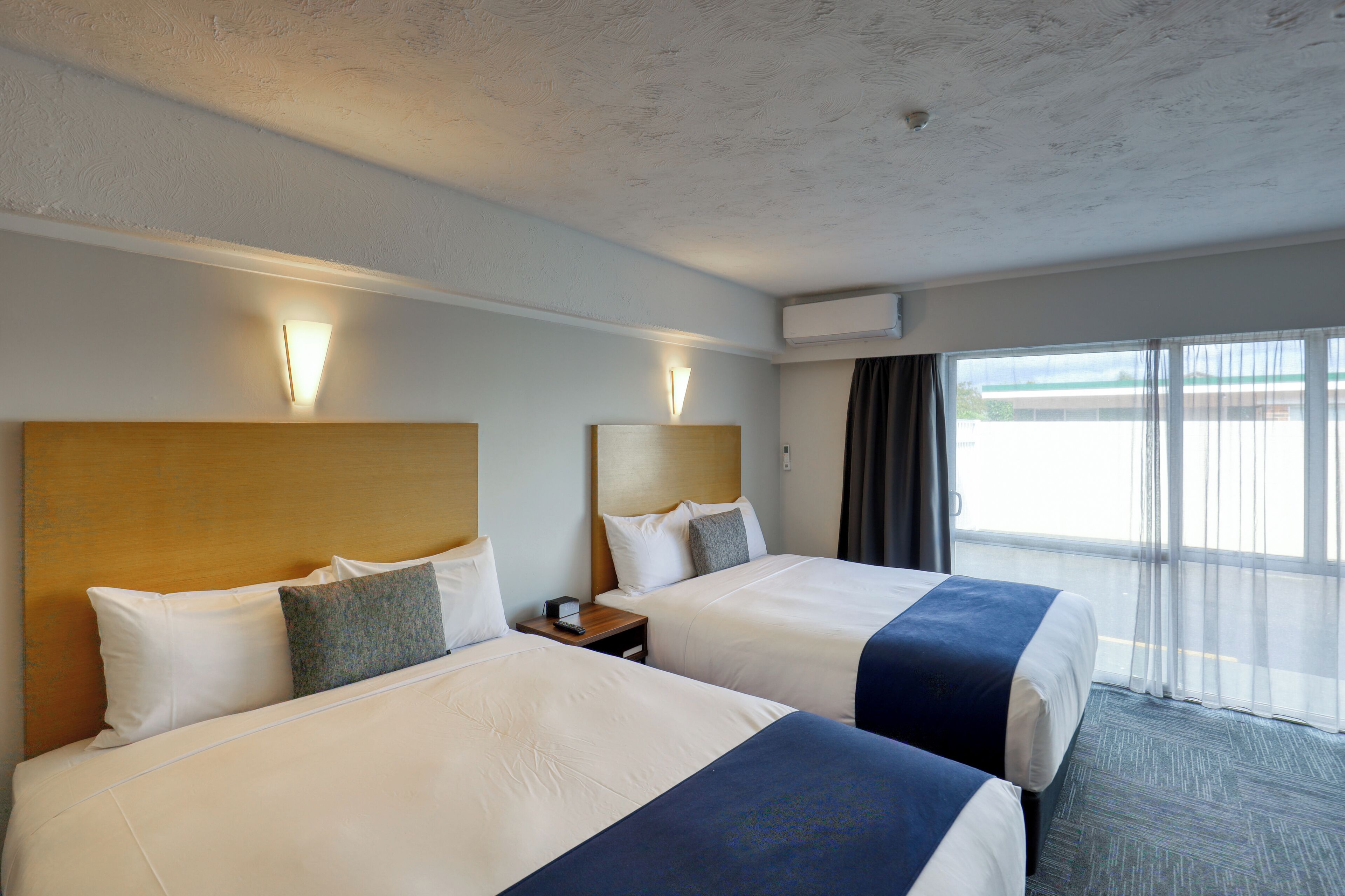 A hotel room with two beds, a nightstand, a lamp, and a large window with curtains.