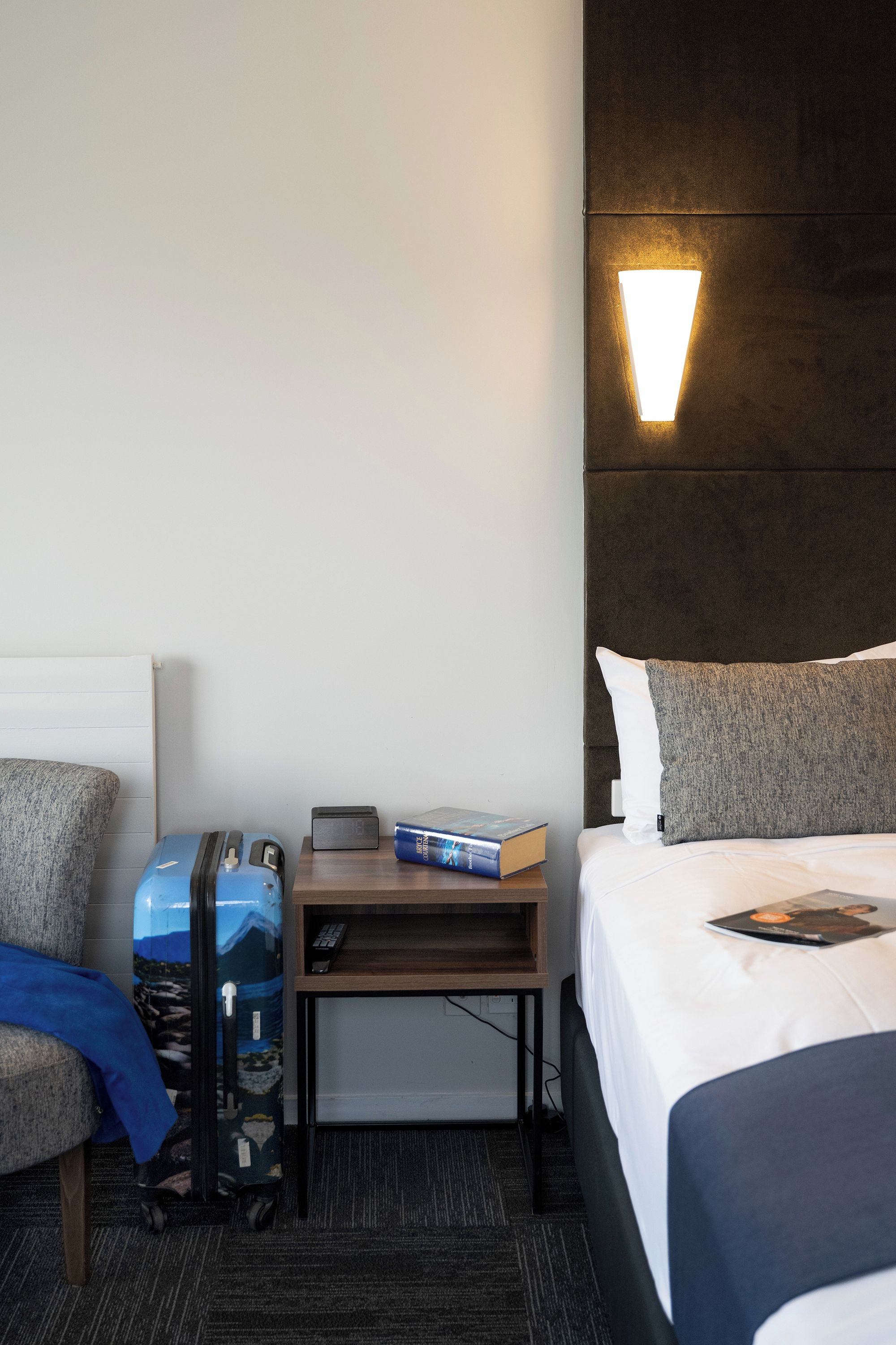 A hotel room with a bed, a bedside table with a lamp, a suitcase, and a book.