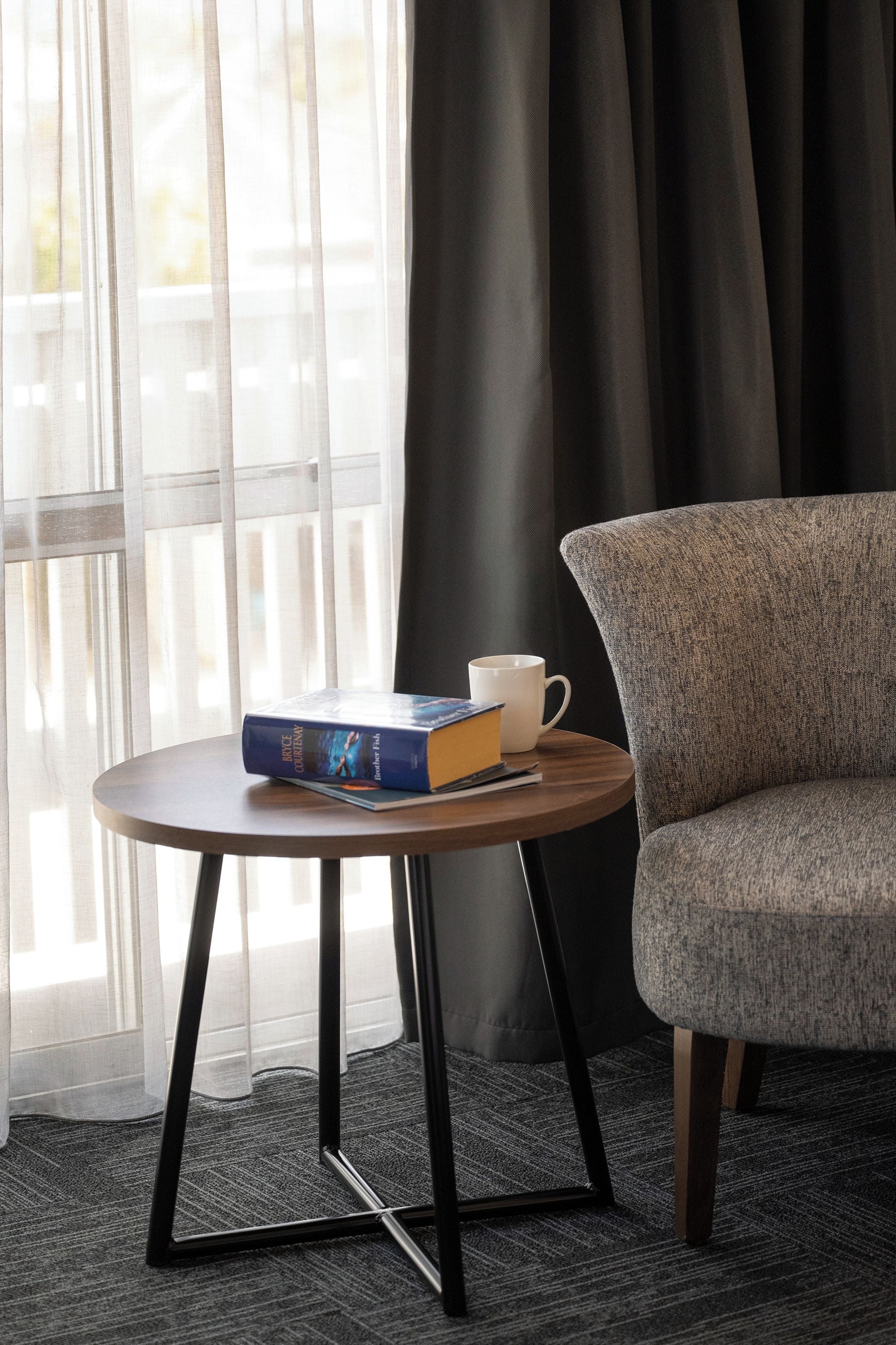 A room with a round table, a chair, a book, a mug, and a window with curtains.