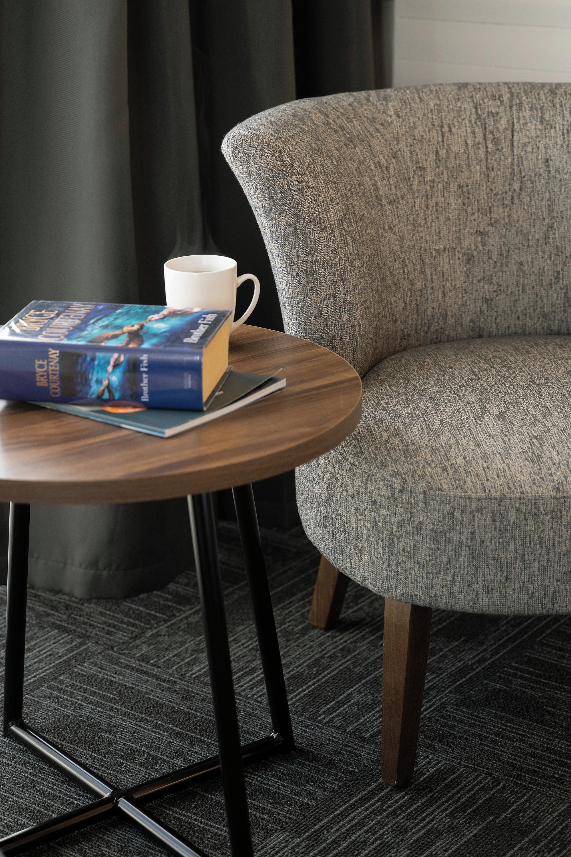 Une table ronde en bois, sur laquelle sont posés un livre et une tasse blanche, est placée devant un fauteuil à revêtement gris.