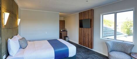A modern hotel room with a large bed, a TV mounted on the wall, a chair, and a window with a view of the outside.