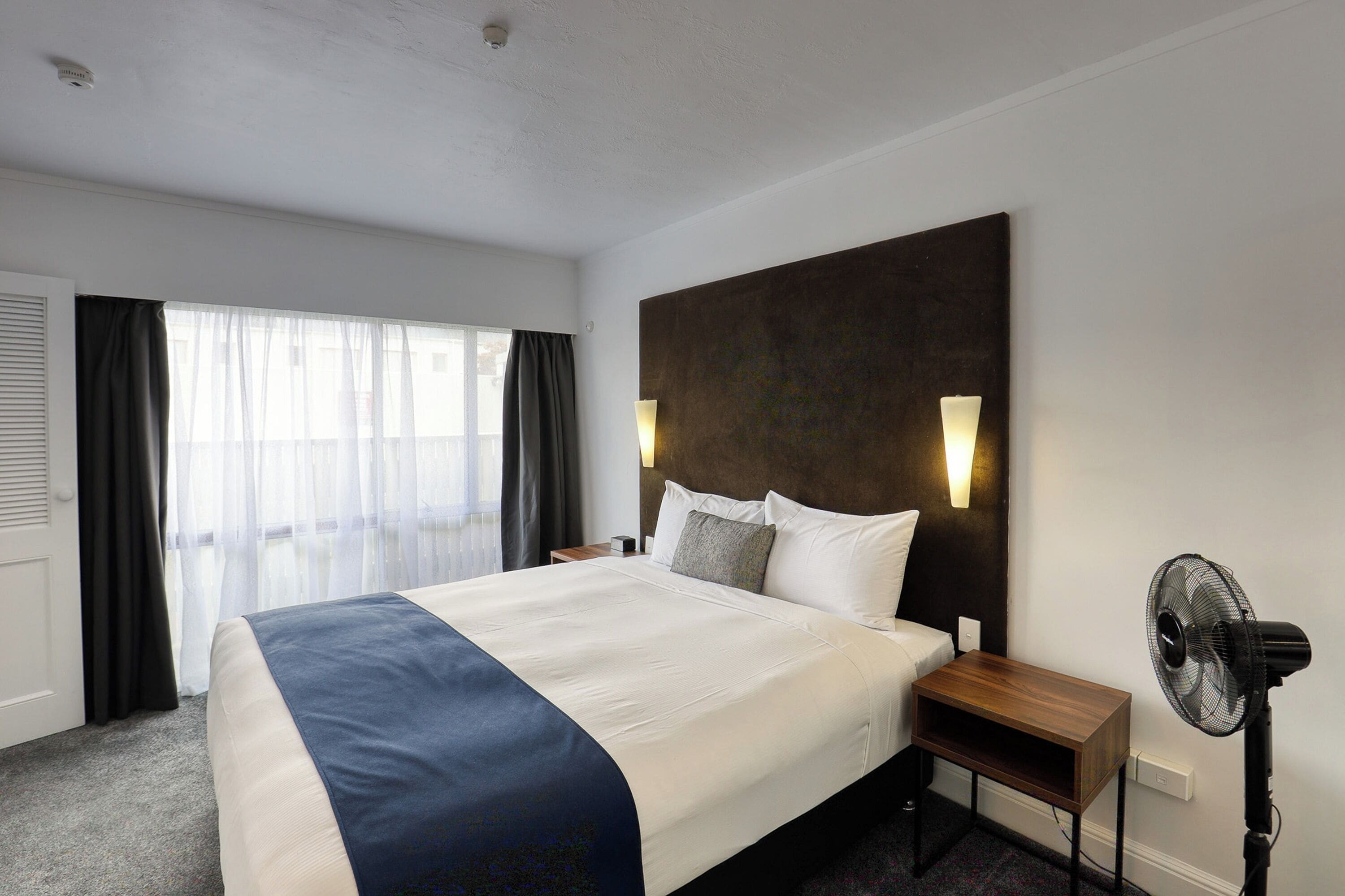 A hotel room with a large bed, a bedside table, a standing fan, and a window with curtains.