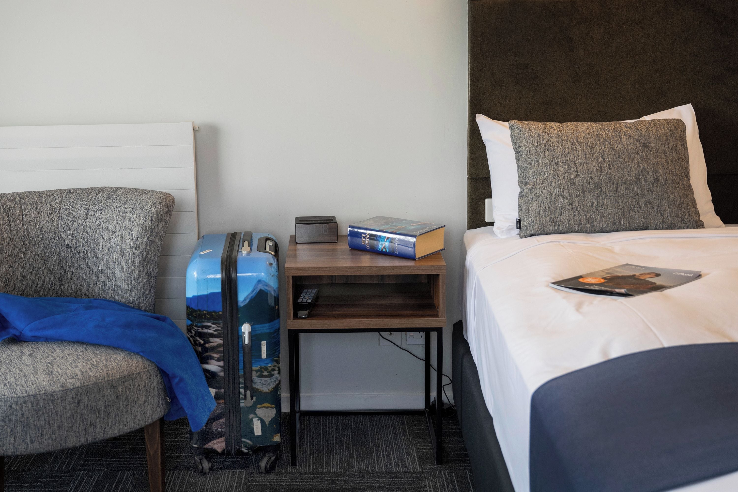 Habitación de hotel con una cama, una silla, una mesita con un libro y un control remoto, y una maleta.
