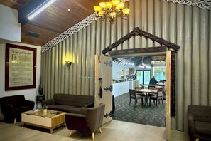 A cozy interior with a wooden ceiling, a chandelier, a framed document on the wall, and a dining area visible through an open doorway.