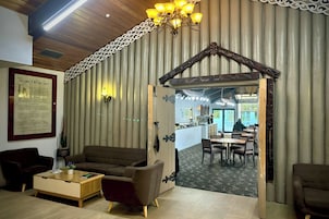 A cozy interior with a wooden ceiling, a chandelier, a framed document on the wall, and a dining area visible through an open doorway.
