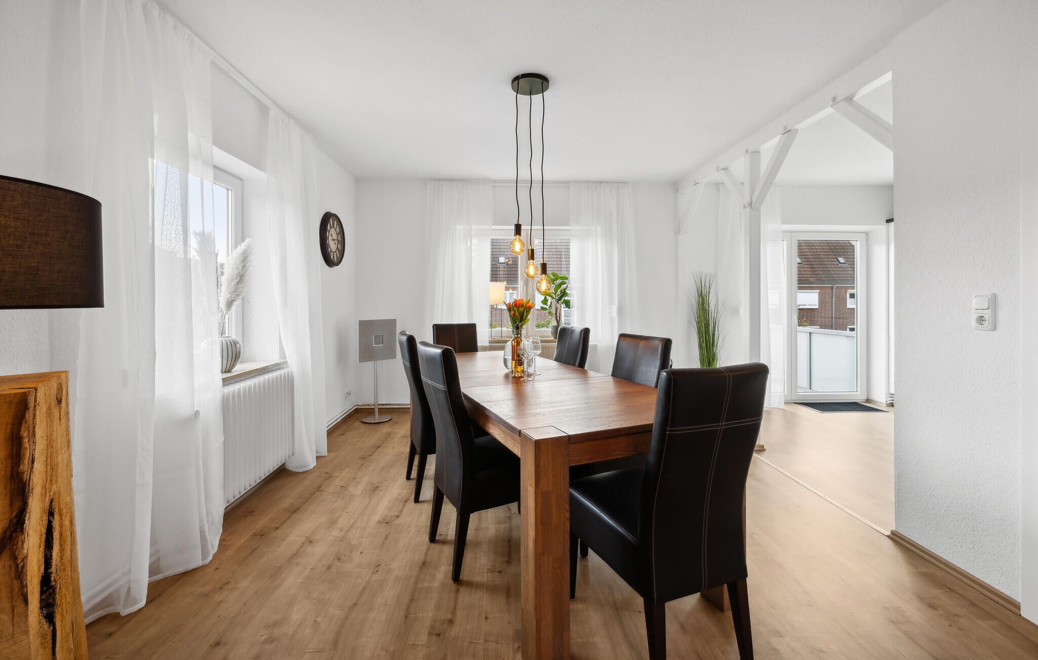 Un comedor con una mesa de madera, sillas negras, una lámpara colgante y un ventanal con cortinas traslúcidas.