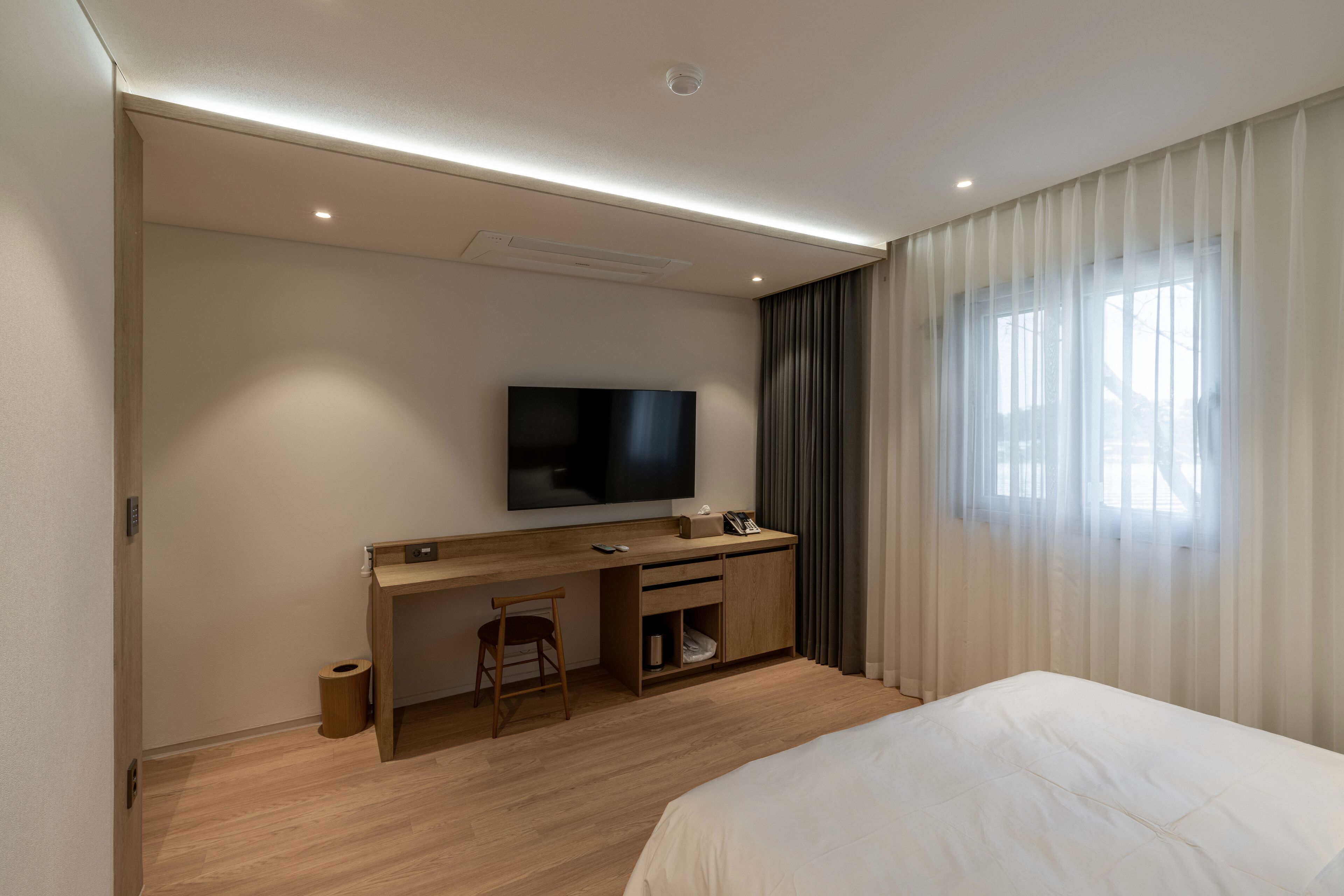 A modern hotel room with a wooden desk, a flat-screen TV, a bed, and a large window with sheer curtains.