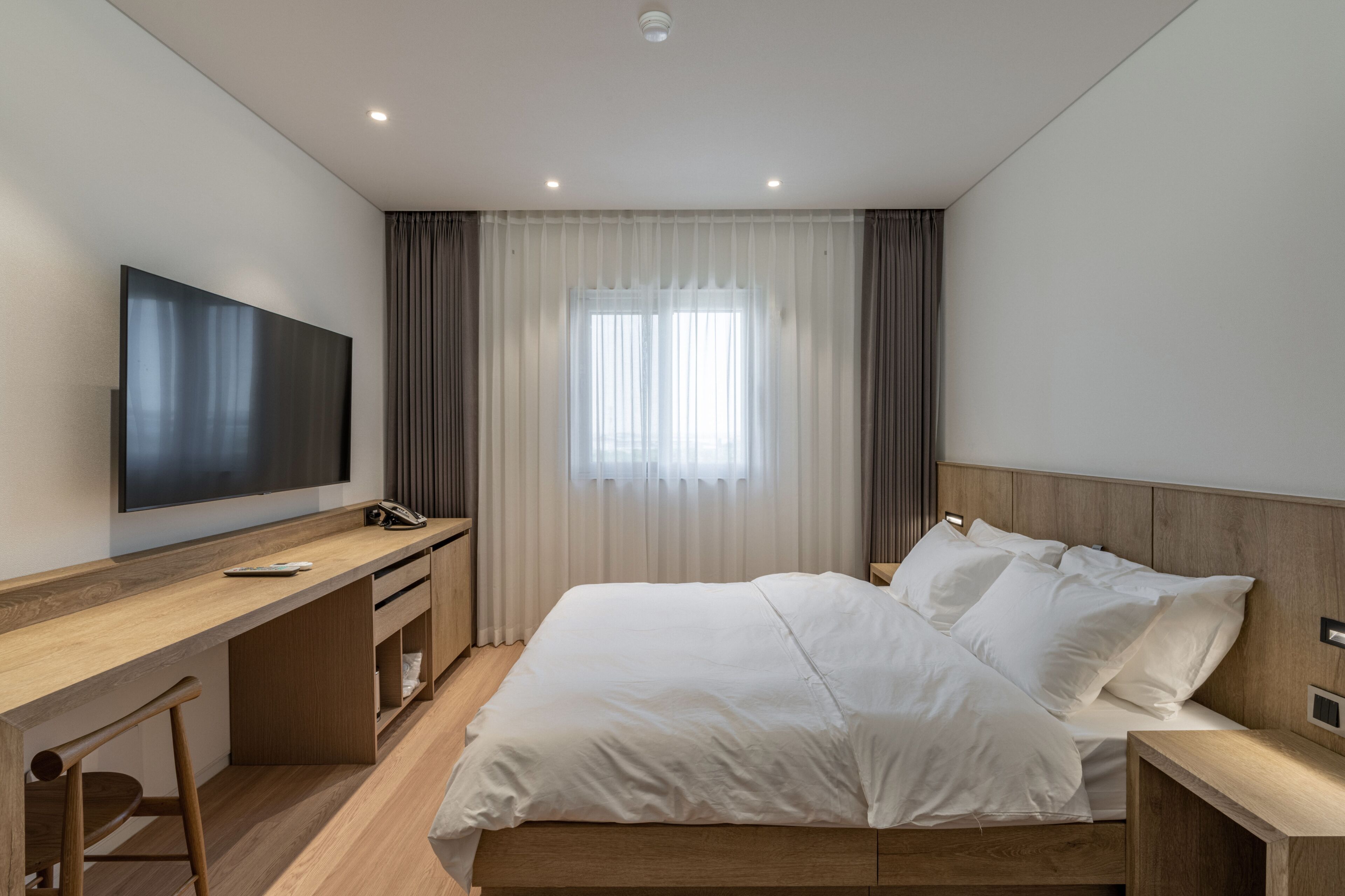 A modern hotel room with a large bed, a desk with a chair, a flat-screen TV, and a window with curtains.