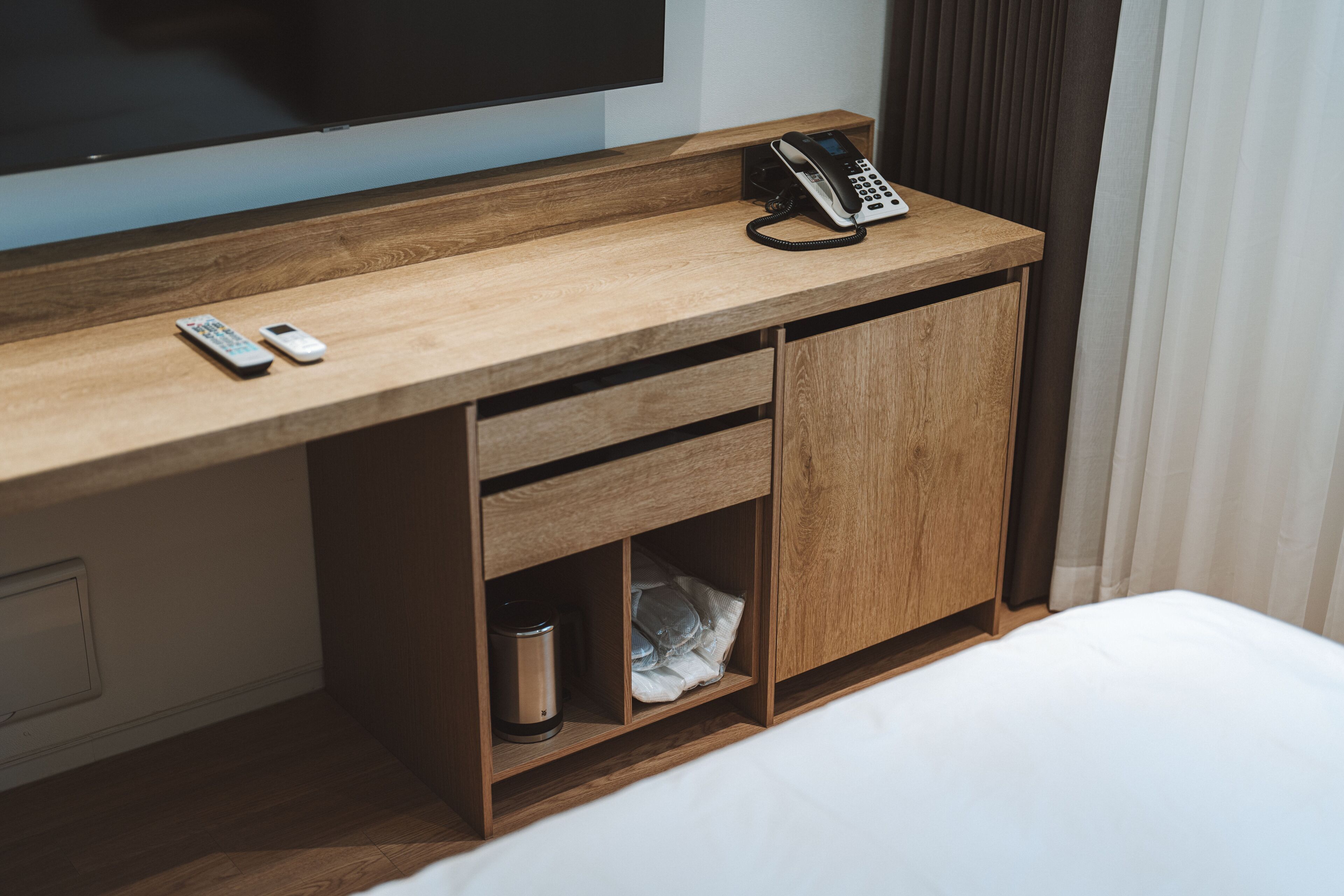 A wooden desk with a telephone, a remote control, and a trash bin.