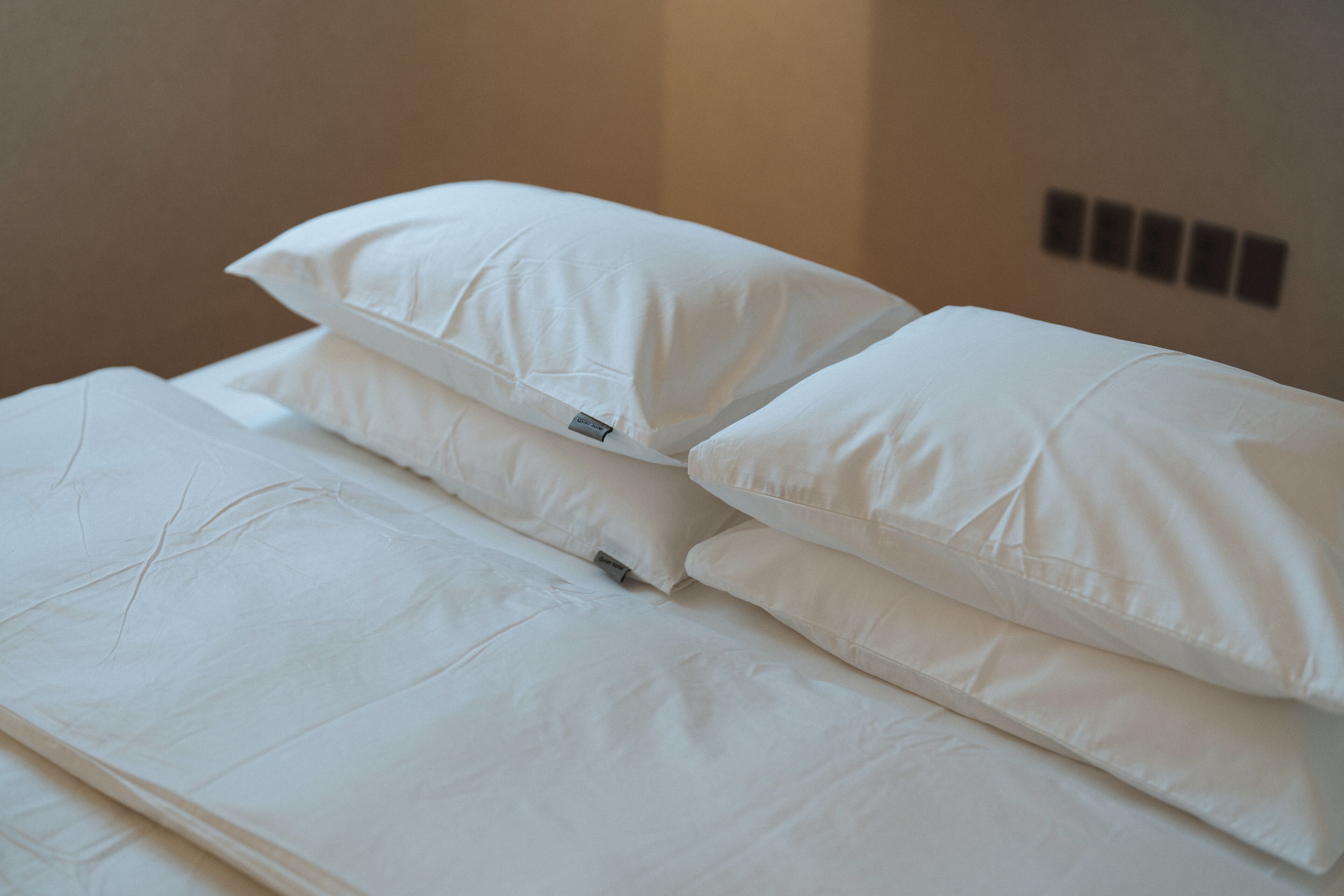 A neatly made bed with white linens and pillows, a visible tag on one pillow, and a wall with a power outlet and a switchboard.