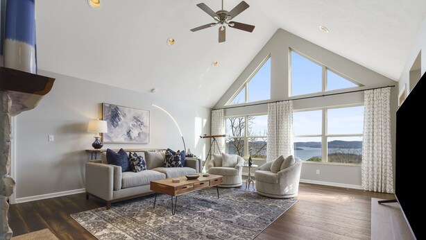 A spacious living room with a high ceiling, large windows, a ceiling fan, and a fireplace.