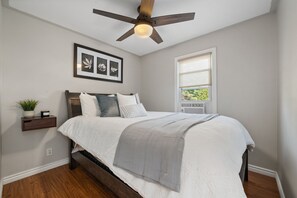 A bedroom with a bed, a ceiling fan, a window with blinds, a wall-mounted shelf with a plant, and framed artwork.