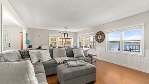 A living room with a gray sectional sofa, a wooden floor, a clock on the wall, and a view of the outdoors. - Barefoot Beach on Pigeon Lake  (Bobcaygeon)