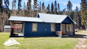 Eine blaue Holzhütte mit Metall-Dach, einer Veranda und einer kleinen Terrasse, umgeben von hohen Bäumen und blauem Himmel.