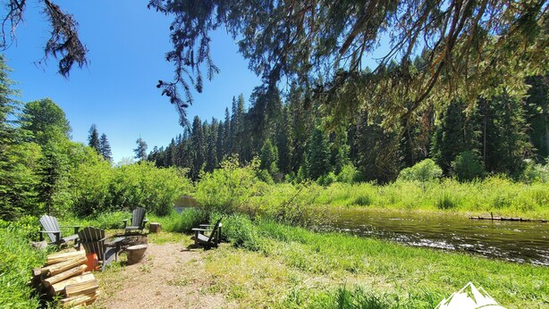 Ein Campingplatz mit Adirondack-Stühlen und einem Picknicktisch in der Nähe eines Flusses, umgeben von dichtem Wald.
