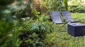 Zwei Lounge-Sessel und ein kleiner Tisch in einem üppigen Garten mit dichtem Grün und einem Gebäude im Hintergrund.