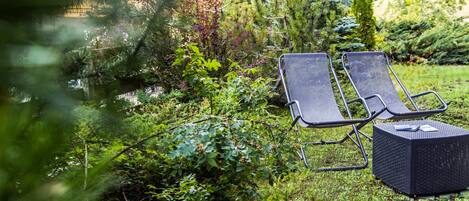 Dos sillones de descanso y una mesita baja en un jardín exuberante con vegetación frondosa y un edificio al fondo.