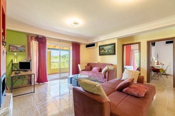 A living room with a sofa, a glass coffee table, a TV, and a balcony with curtains.