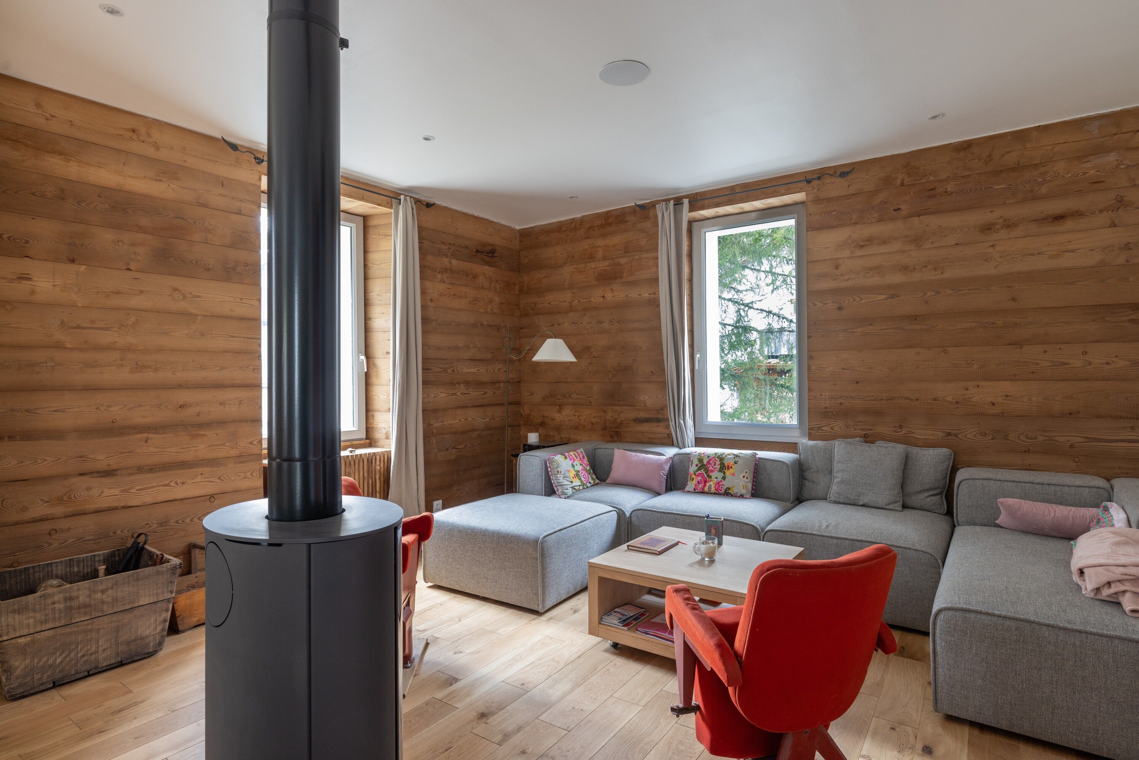Een gezellige woonkamer met een houtkachel, een grijze bank, een rode fauteuil en een raam met gordijnen.