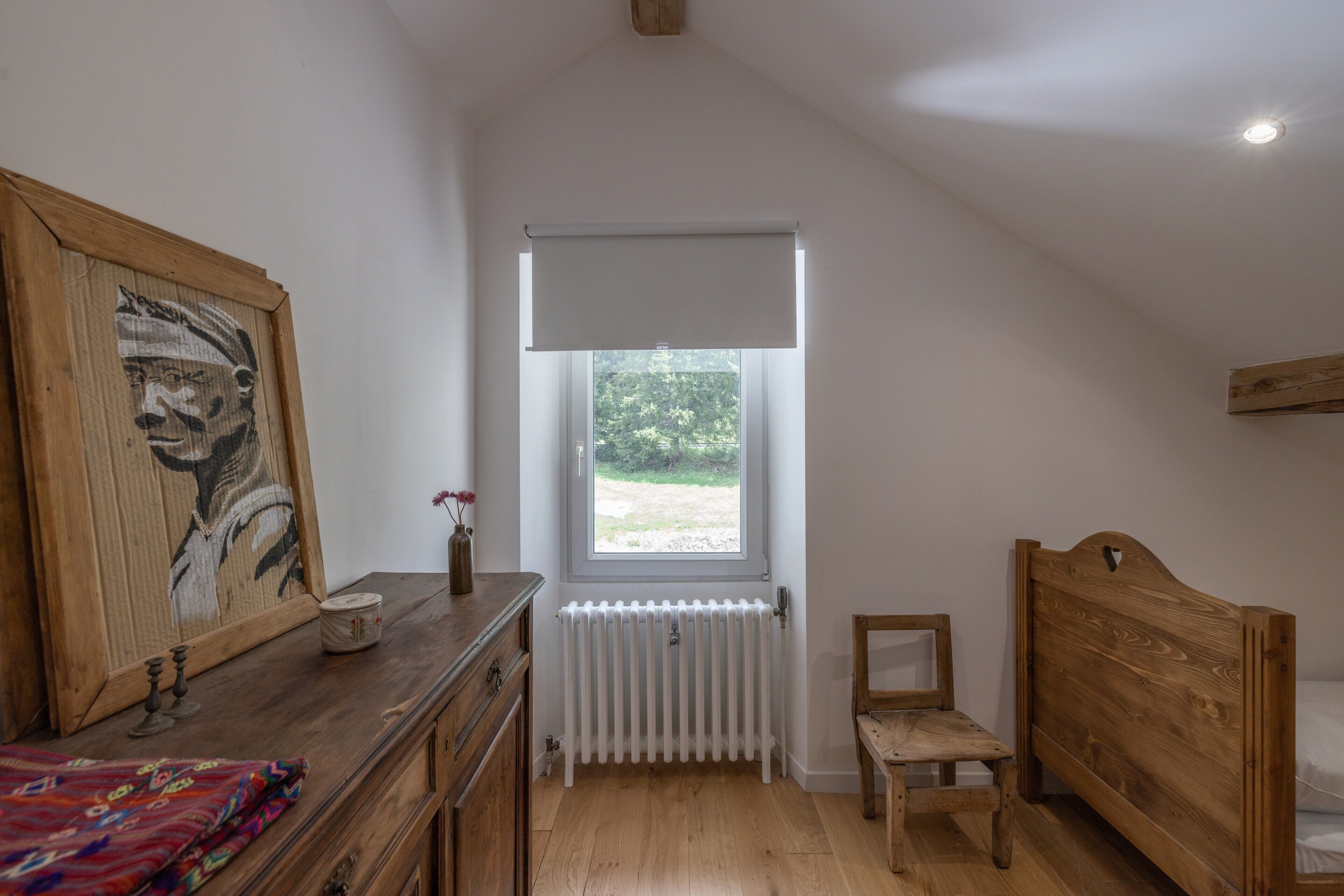 A bedroom with a wooden bed, a radiator, a window with a view of greenery, and a wooden dresser with a mirror and a vase.