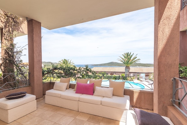 A covered outdoor seating area with a sofa and ottoman, a pool, and a view of hills and a clear sky.