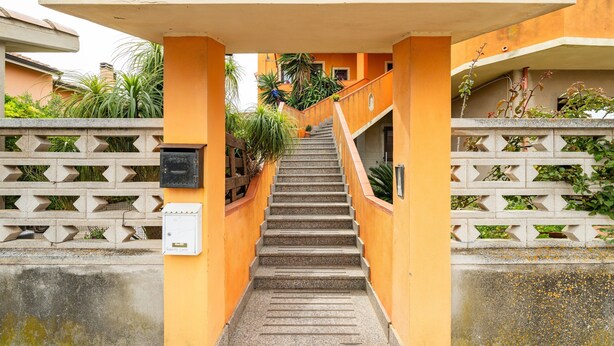 A staircase leading to a building with a yellow facade, a concrete wall with a geometric pattern, and some greenery.