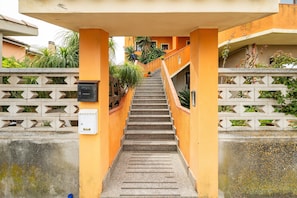 Un escalier menant à un bâtiment à la façade jaune, un mur en béton orné d’un motif géométrique et quelques plantes vertes.