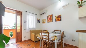 A dining area with a table, chairs, and a sofa. There is a window with curtains, a wall-mounted air conditioner, and a potted plant.