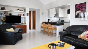 Uma sala de estar moderna com televisão, sofás, uma mesa de centro e uma cozinha ao fundo.