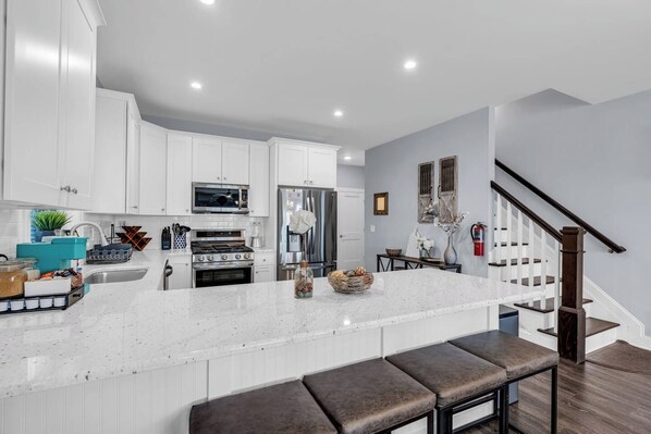 A modern kitchen with white cabinetry, a large island with bar stools, stainless steel appliances, and a staircase with wooden steps.