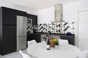 Een moderne keuken met een roestvrijstalen koelkast, magnetron en fornuis. Er staat een eettafel met stoelen, een fles wijn en een potplant.